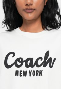 Camiseta de algodón blanca con letras negras en negrita que dicen "Coach NEW YORK" en una fuente de escritura divertida. Textura suave y diseño de cuello redondo.