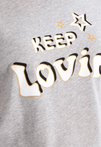 Camiseta de algodón gris con el texto "SIGUE AMANDO" en letras gruesas y bordadas. Acentos de estrellas notables en amarillo y contornos negros.