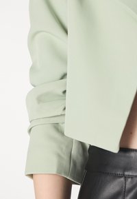 Chaqueta corta verde claro con cuello doblado y mangas remangadas, combinada con una falda de cuero negra de talle alto. Textura suave.