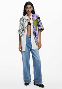 Camicia a fantasia floreale bianca e viola abbinata a jeans a gamba larga blu chiaro. La camicia presenta grafiche audaci e una vestibilità rilassata.