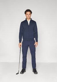 Suéter azul-marinho com fecho, camisa de gola creme, calças azuis-marinho, sapatos de golfe pretos. Em pé com um taco de golfe contra um fundo neutro.