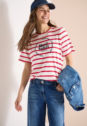 Uśmiechnięta kobieta w czerwono-białej koszulce w paski z napisem, niebieskich dżinsach, niebieskiej czapce, trzymająca kurtkę dżinsową w lewej ręce.