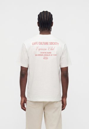Personne aux cheveux tressés portant un t-shirt blanc avec un texte rouge listant une adresse de café à Brooklyn dans le dos, debout devant un fond uni.