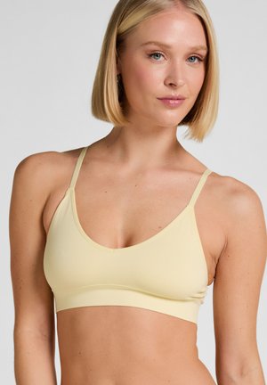 Blonde Frau mit Bob-Frisur, die einen hellgelben nahtlosen Bralette mit dünnen Trägern vor einem grauen Hintergrund trägt.