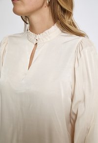 Beige Bluse mit hohem, rüschigem Kragen, Schlüssel-Loch-Ausschnitt am Hals und Puffärmeln. Glattes Material mit leichtem Glanz.