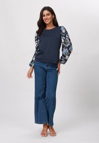 Marineblå topp med blomsterprydede langermede, kombinert med vide blå jeans og nude sandaler med stropper, med en avslappet passform og glatt tekstur.
