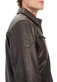 Bruine leren jas met een klassieke kraag, zijzak met rits en eenembossed logo-detail op de linkermouw. Glad textuur en getailleerde pasvorm.