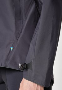 Donkergrijze waterdichte jas met een gladde textuur. Beschikt over een blauw en geel embleem en een zwart verstelbaar manchetklem voor een veilige pasvorm.