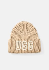 Căciulă tricotată de culoare bej, cu o textură în dungi și o margine îndoită. Prezentă cuvântul "UGG" brodat cu alb pe margine.