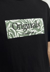 Schwarzes T-Shirt mit einem rechteckigen grünen und weißen Grafikdesign, das den Text "Originals" und "JACK&JONES" in fetten Buchstaben zeigt.