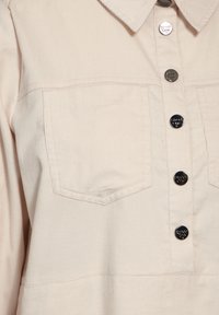 Chemise beige clair en tissu texturé, dotée d'un col, de boutons argentés et de deux poches poitrine. Le design est à la fois structuré et décontracté.