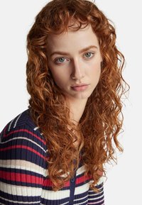 Pletený pruhovaný kardigan v námořnické modré, červené a bílé barvě; ribbovaný povrch a zapínání na knoflíky. Model má kudrnaté červené vlasy a neutrální výraz.