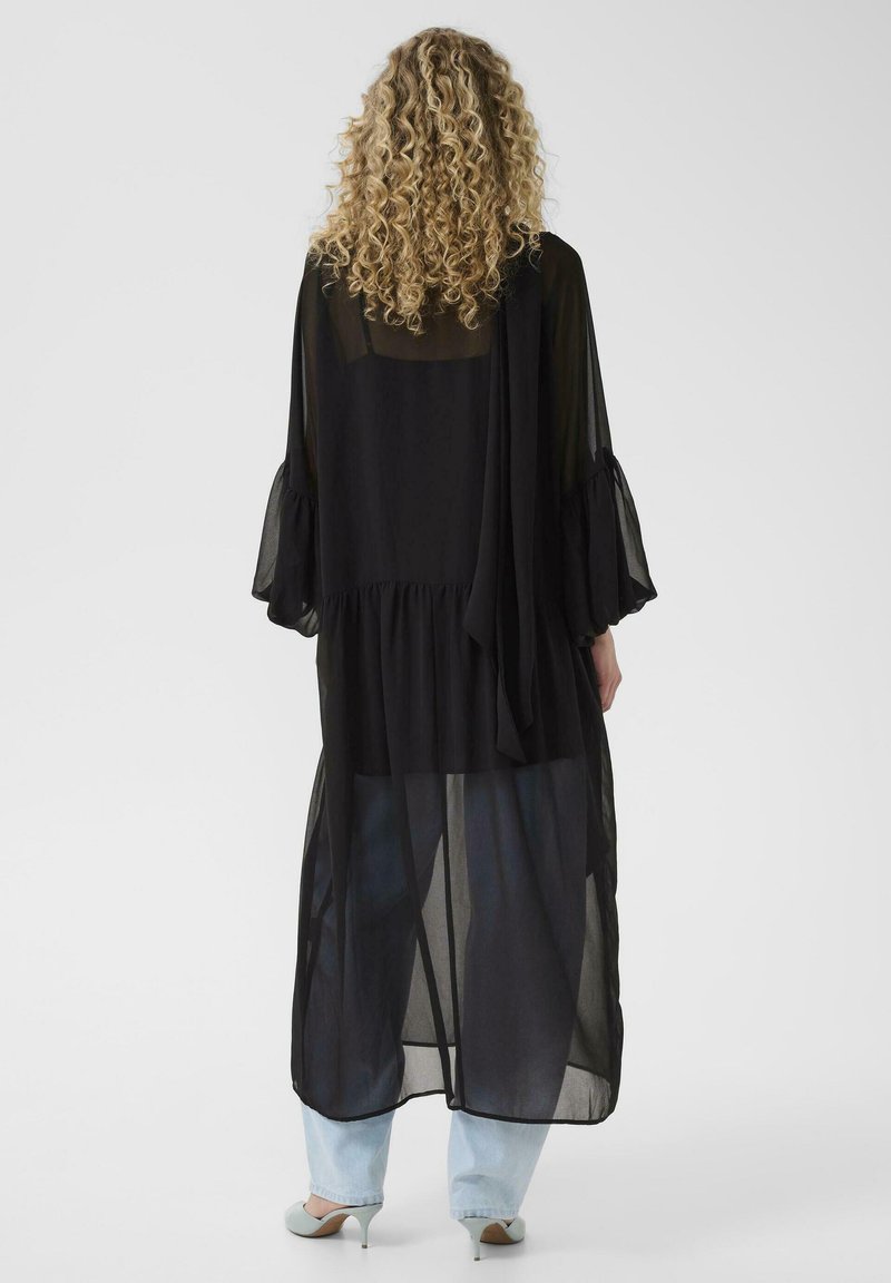 Femme aux cheveux blonds bouclés portant une tunique noire transparente à manches longues sur un jean bleu clair et des escarpins bleu pâle, vue de dos.