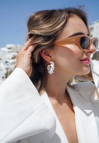 Weiße gedrehte Creolen mit goldenen Akzenten, getragen von einer Person in einem weißen Blazer und braunen Sonnenbrillen vor einem blauen Himmel.