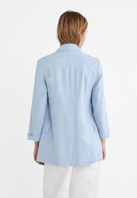 Hellblauer, maßgeschneiderter Blazer mit Kerbkragen, dreiviertellangen Ärmeln und Rückenschlitz-Details. Der Stoff wirkt glatt mit einer dezenten Struktur.