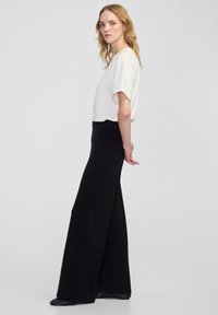 Weißes Kurzarm-Oberteil, kombiniert mit hochschnittigen, schwarzen weiten Hosen. Glatte Stoffe, lässige Passform und subtile Texturen bestimmen das Outfit.