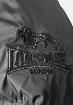 Schwarzer Stoff mit einem gestickten Logo, das einen Löwen zeigt, mit dem Wort "LONSDALE" darüber und "LONDON" darunter, alles in schwarzem Faden.