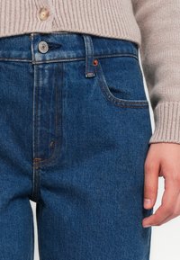 Blauwe denim jeans met een hoge taille, voorzien van koperen klinknagels, voorzakken en een soepele textuur, gecombineerd met een beige gebreid sweater.