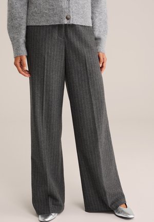 Femme portant un pantalon large à fines rayures grises, un cardigan gris boutonné et des chaussures pointues argentées, debout devant un fond uni.
