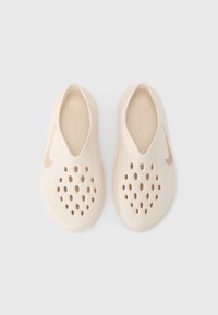 Μπεζ παπούτσια τύπου slip-on κατασκευασμένα από υλικό με υφή, με πολλαπλές στρογγυλές οπές και μικρό λογότυπο Nike στην πλευρά. Ομαλός, στρογγυλεμένος σχεδιασμός.