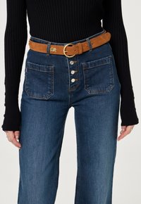 Hoge taille donkerdenim jeans met een sluiting van vijf knopen, twee zijzakken en een bruine suede riem met een ronde gouden gesp.