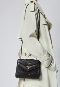 Borsa a tracolla in pelle nera con motivo a zig zag, accentuata da un logo dorato e una tracolla a catena, indossata sopra un trench coat chiaro.