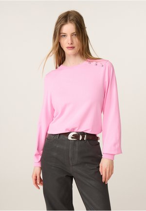 Jonge vrouw die een lichtroze top met lange mouwen draagt met knopen op de schouder en donkere hoge taillebroeken met een zilveren gesp.