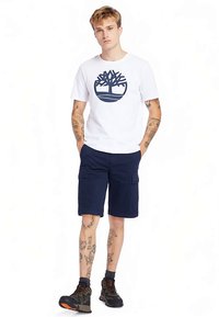 Bílé tričko s námořnicky modrým logem stromu, doplněné o námořnicky modré cargo kraťasy. Model má na nohou černo-šedé tenisky. Neformální outfit, viditelné tetování.