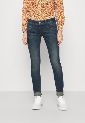 Ciemnoniebieskie, dopasowane jeansy z podwiniętymi nogawkami noszone z białymi trampkami i pomarańczową, kwiatową koszulą z długim rękawem na stojącej osobie.