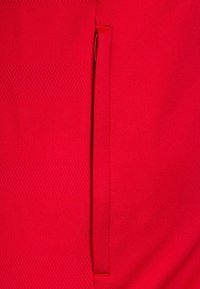 Roter Stoff mit einer glatten Textur und einem vertikalen Streifenmuster. Enthält eine kleine, versteckte Tasche in der linken Naht.