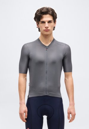 Tânăr purtând un tricou de ciclism gri, cu mâneci scurte și ajustat, și pantaloni scurți de ciclism căptușiți, bleumarin, stând în picioare pe un fundal simplu.