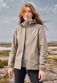 Red-haired woman wearing a beige zip-up jacket and green top stands outdoors on rocky terrain with cloudy sky and water in the background.