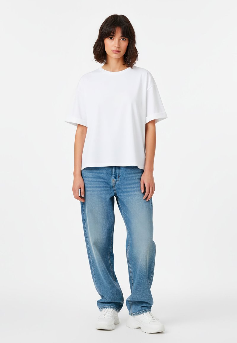 T-shirt bianca oversize a maniche corte abbinata a jeans azzurri morbidi e scarpe da ginnastica bianche. Design semplice, texture liscia e taglio classico.