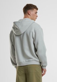 Hellgrauer Hoodie mit lockerem Schnitt, ausgestattet mit einer Kängurutasche, gerippten Bündchen und einer Kapuze mit Kordelzug, kombiniert mit olivgrünen Hosen.