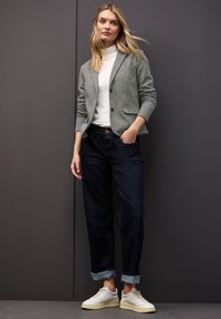 Grijze wolmix blazer met puntige revers, witte coltrui, donkerdenimjeans en crèmekleurige sneakers; casual pasvorm met opgerolde mouwen.