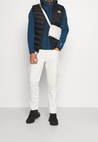 Czarna pikowana kamizelka na niebieskiej bluzie z suwakiem, w połączeniu z białymi spodniami i czarnymi trampkami. Nosi małą torebkę na ramię w jasnoszarym kolorze.