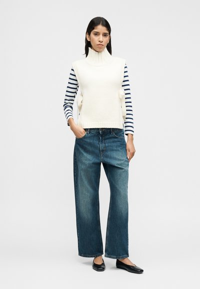 Ελεφαντόδοντο γιλέκο ζιβάγκο με ριγέ μακρυμάνικη μπλούζα, σε συνδυασμό με τζιν παντελόνι φαρδιάς γραμμής και μαύρα επίπεδα παπούτσια. Απλός, μινιμαλιστικός σχεδιασμός.