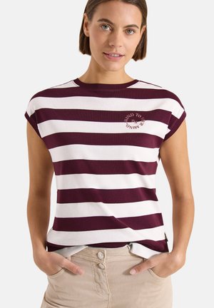 Donna che indossa una maglietta a maniche corte bianca e viola scuro a righe orizzontali, con le mani nelle tasche di pantaloni beige.