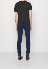 Černé bavlněné tričko v kombinaci s modrými kalhotami na míru; model otočený zády, ukazuje slim fit a texturovanou tkaninu. Černé kotníkové boty.