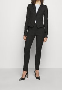 Černý přizpůsobený blejzr s ramenními vycpávkami, zapínání na jeden knoflík a přiléhavé černé kalhoty. Úzký střih, hladká tkanina a špičaté černé podpatky.