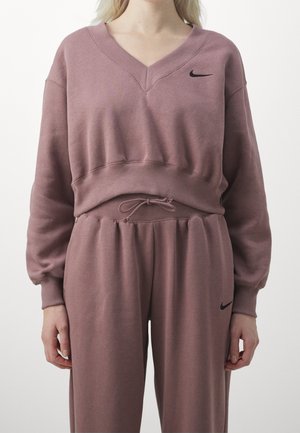 Kobieta ubrana w liliową bluzę Nike i pasujące spodnie dresowe z regulowanym sznurkiem w talii oraz czarnymi logo Nike na piersi i udzie.