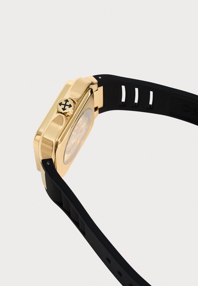 Boîtier de montre carré doré avec bracelet en silicone noir, avec une couronne présentant un détail de logo noir à quatre flèches sur un fond clair uni.
