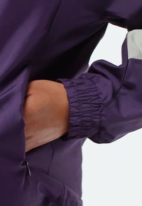 Hand inserted into the elastic cuff pocket of a purple jacket with a white stripe detail on the sleeve, against a white background.