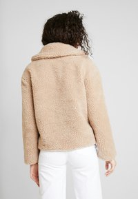 Osoba s kudrnatými vlasy má na sobě měkký, béžový, teddy kabát, který je otočená zády k jednoduchému šedému pozadí. Kabát má široký límec a volný střih.