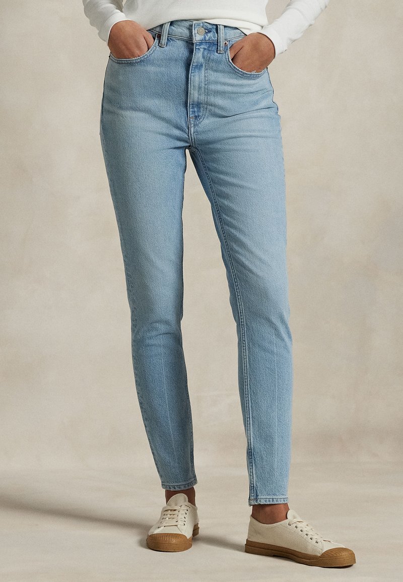 Persoon draagt lichtblauwe skinny jeans met een hoge taille, handen in de zakken en witte sneakers met bruine zolen op een neutrale achtergrond.