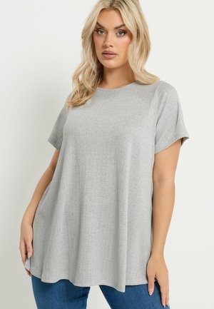 Donna bionda con indosso una maglia grigia a maniche corte e larghe e jeans blu, in piedi davanti a uno sfondo chiaro e semplice.