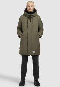 Olivenfarvet parka med stor hætte, frontlommer og subtile syninger. Sættes sammen med sorte jeans og sorte støvler.
