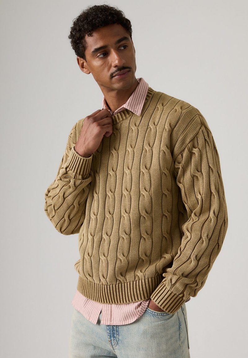 Man adjusting collar wearing beige cable-knit sweater over pink striped shirt and light blue jeans against plain background.