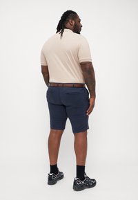 Beige polo shirt med korte ærmer, marineblå shorts, brun læderbælte og sorte sportsko. Glat stof og skræddersyet pasform.