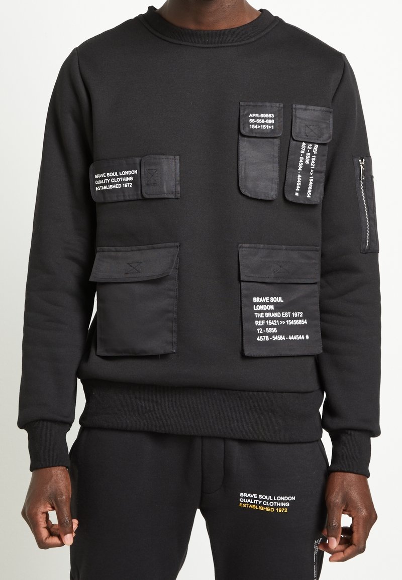 Černá mikina s několika texturovanými kapsami se záplatami a potiskem etiket. Žebrovaný výstřih a manžety. Kapsy mají matný povrch.